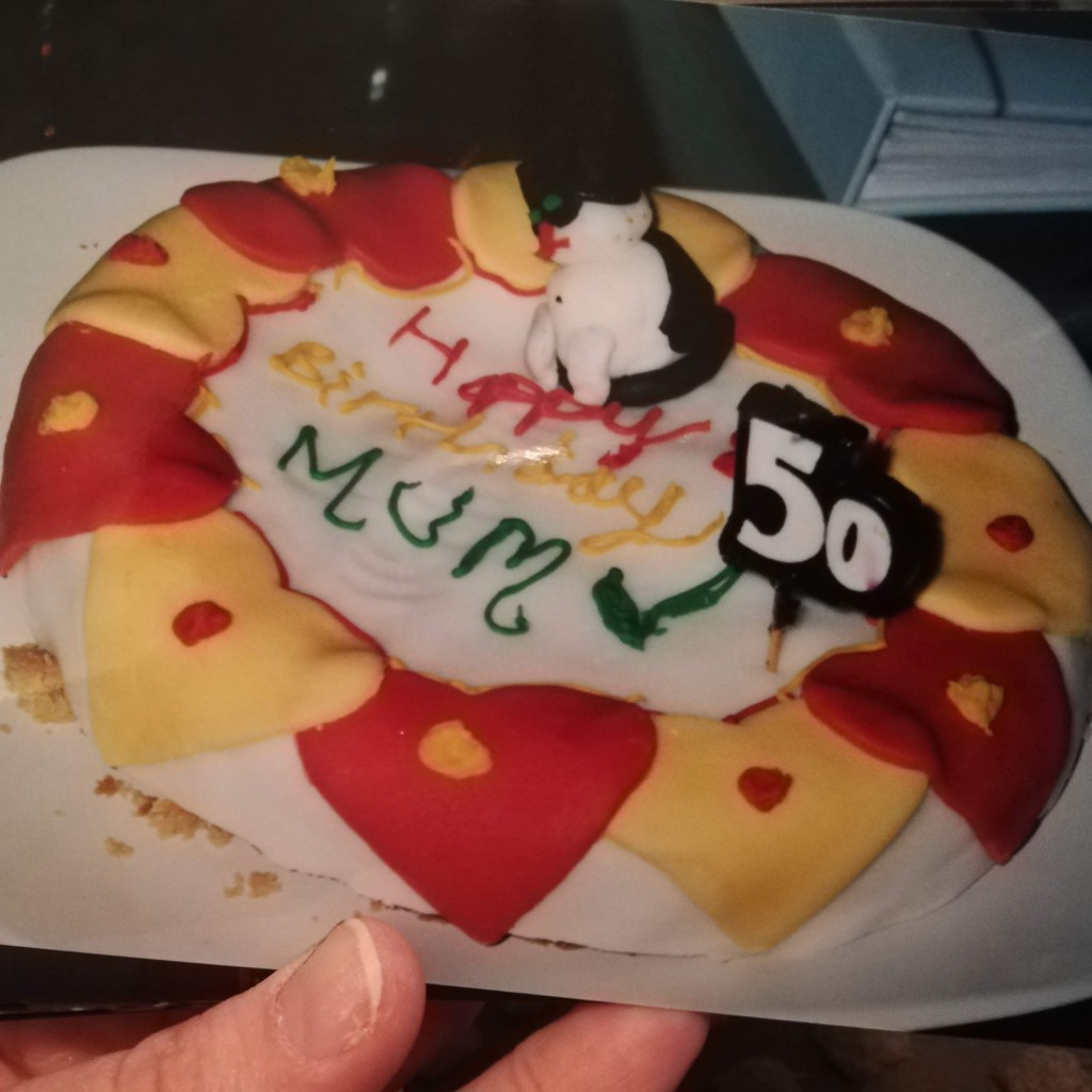 A terribly decorated flat cake with white royal icing on top and red and yellow fondant hearts around the edge. In the middle sits a fondant black and white cat and writing that says "happy birthday Mum"