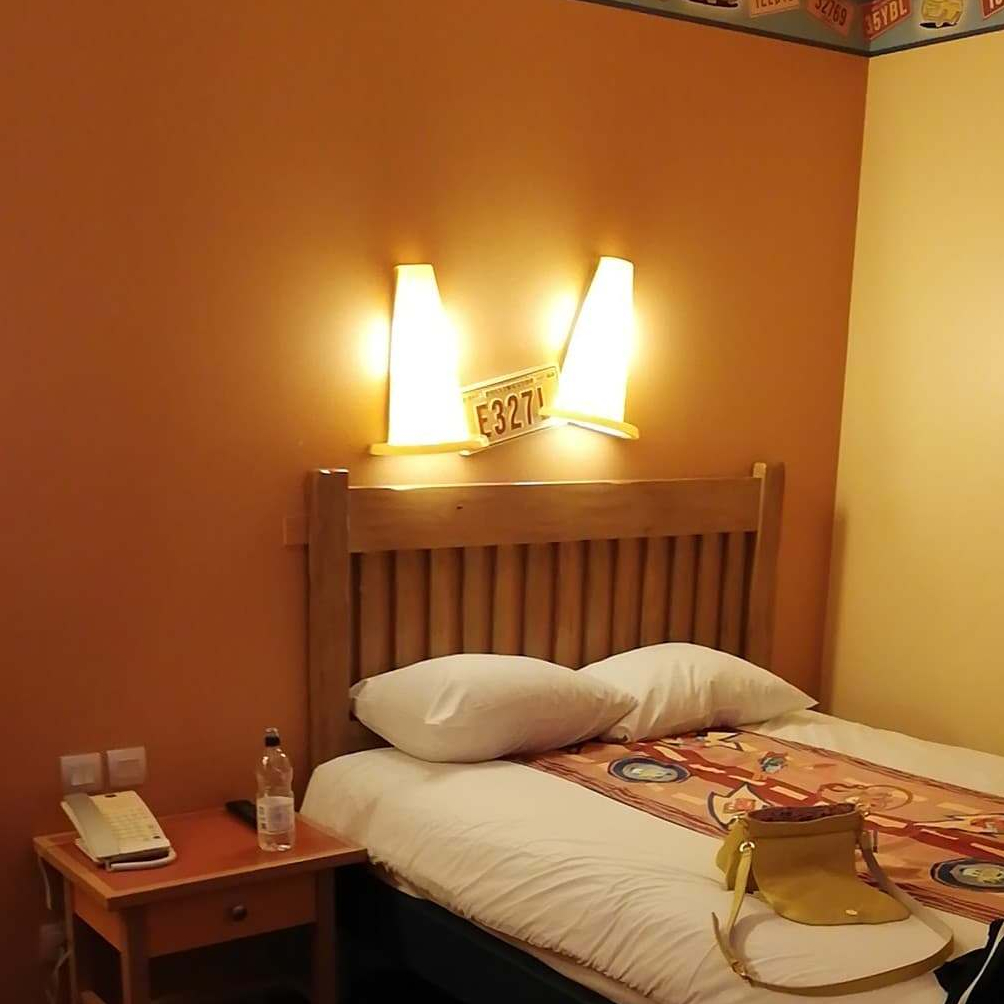 My room at the Disney Santa Fe Hotel - my bed is against orange walls with traffic cone shaped lights. There is a white duvet and pillows with a Cars themed runner down the middle.