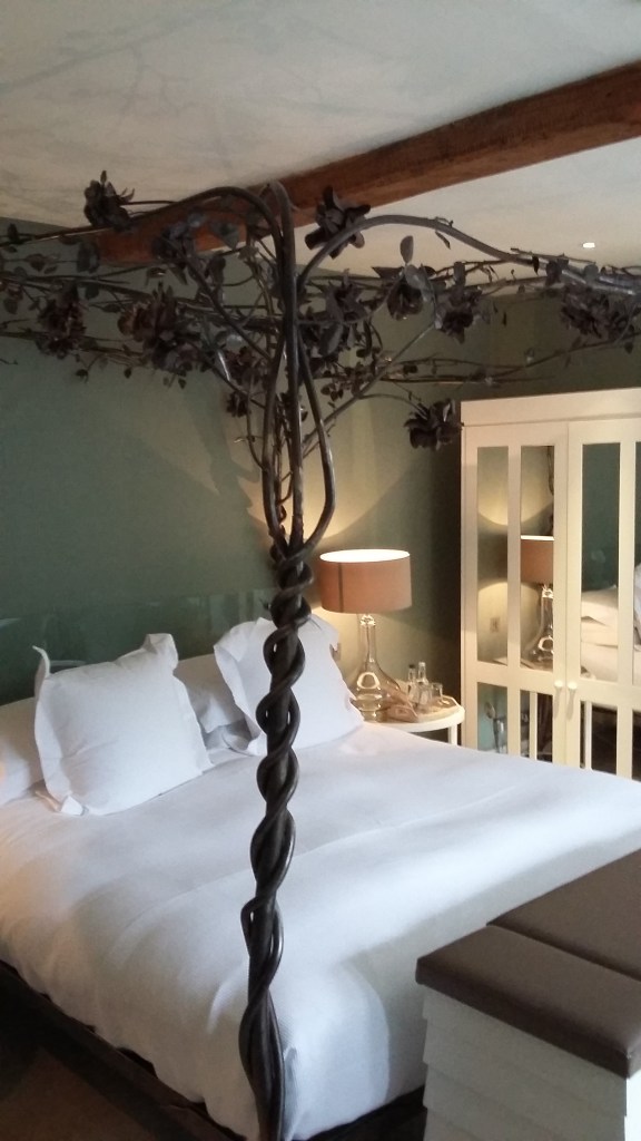 A photo of the bedroom at Barnsley House Hotel. The four-poster bed has a wire canopy decorated with metal leaves and the bed is covered in fresh white sheets with huge pillows.