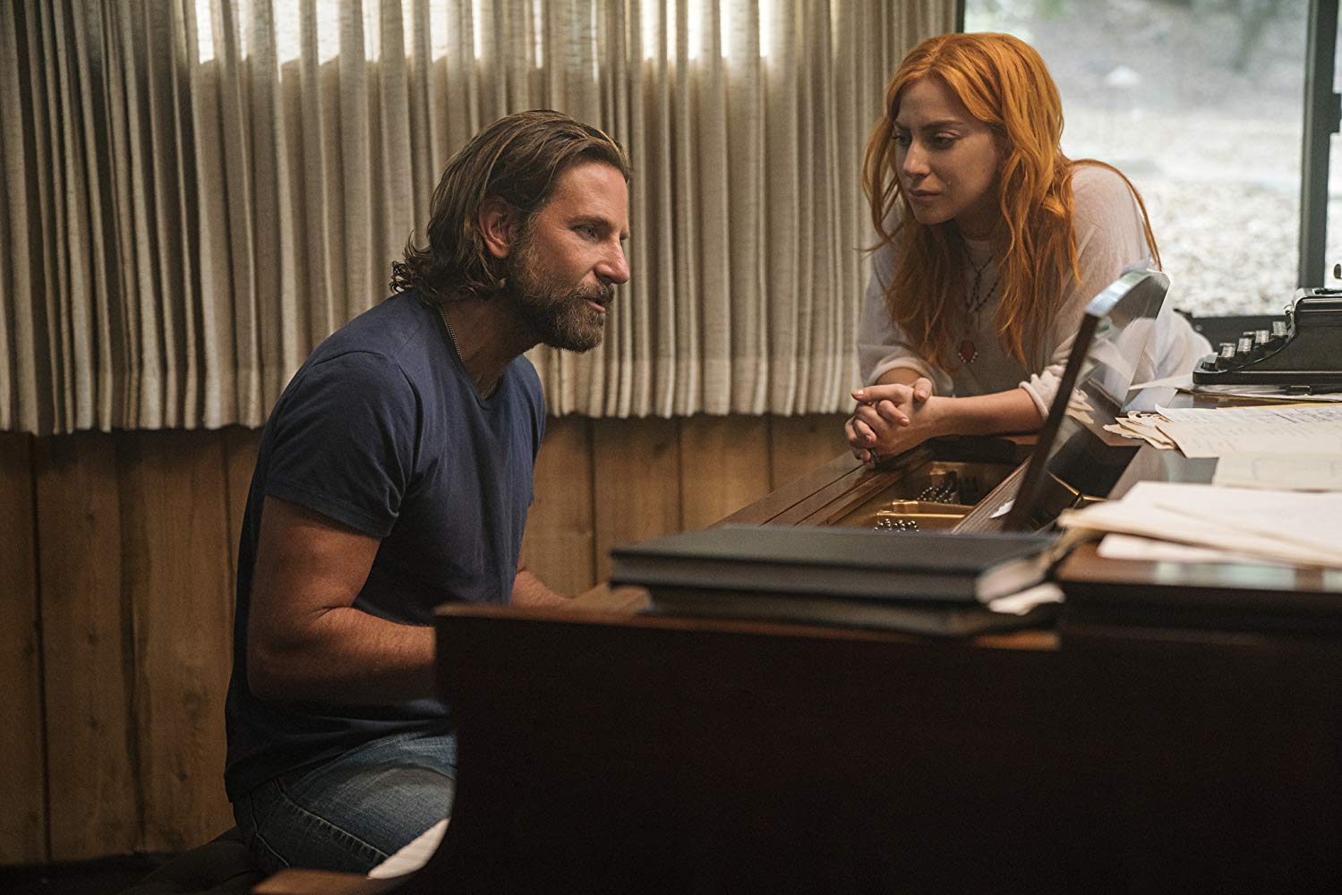Still from A Star is Born - Bradley Cooper's character, Jackson Maine, sits at a piano, singing. Lady Gaga's character, Ally, leans against the piano, looking visibly moved.