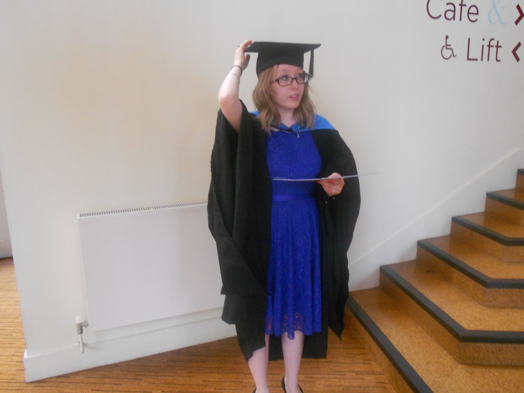 Me after my graduation ceremony wearing the cap and gown. I'm holding my certificate while trying to keep the cap on my head.