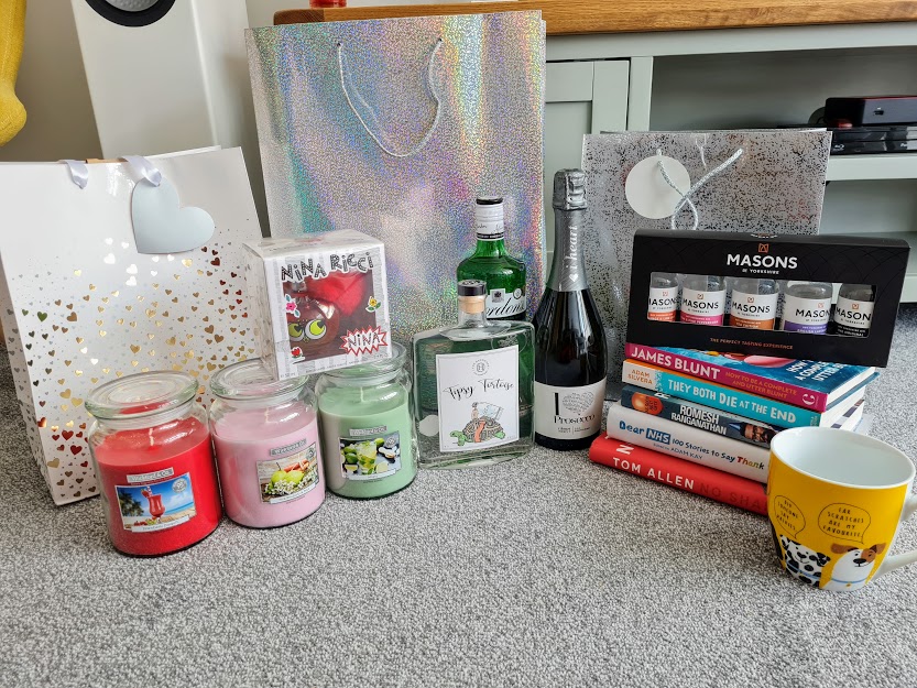 My birthday gifts - three scented candles, a bottle of Nina Ricci perfume, two bottles of gin, a bottle of prosecco, a tasting selection of gin, a stack of books and a yellow mug with dogs on.