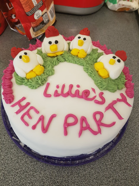 A white cake decorated with royal icing and fondant chickens. It says "Lillie's Hen Party."