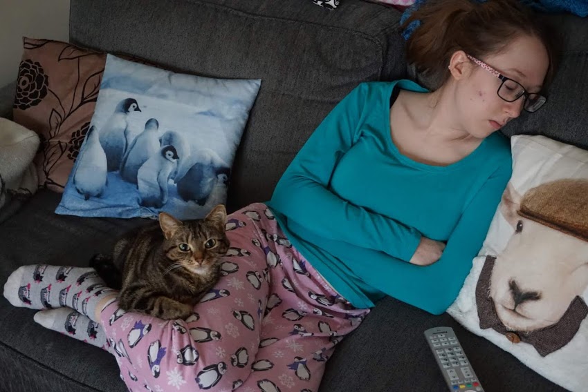 Photo of me in my pyjamas, looking very worse for wear, asleep on the sofa. Tilly is nestled in behind my knees looking bright eyed and bushy tailled.