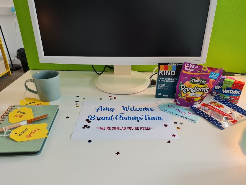 My desk at my new job. There is a sign in the middle that's surrounded by confetti and says "Amy - welcome to the Brand Comms Team - We're so glad you're here!" On the left, there's a mug, a notebook and a fancy pen, and on the right is a box of Kind bars, a packet of Rowntrees Randoms, two boxes of Nerds, and a bag of Jelly Belly jelly beans. 