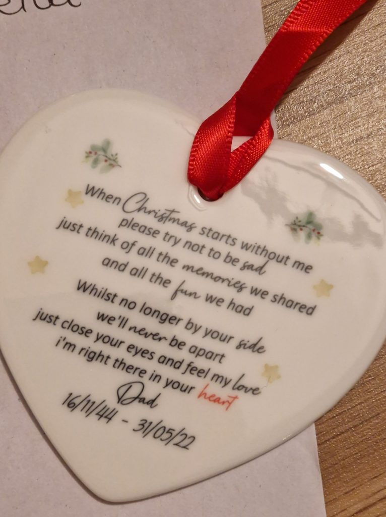 A porcelain, heart shaped Christmas decoration with a red ribbon at the top. It reads "When Christmas starts without me, please try not to be sad, just think of all the memories we shared and all the fun we had. Whilst no longer by your side, we'll never be apart. Just close your eyes and feel my love, I'm right there in your heart. Dad." At the bottom it reads 16/11/44 - 31/05/2022.
